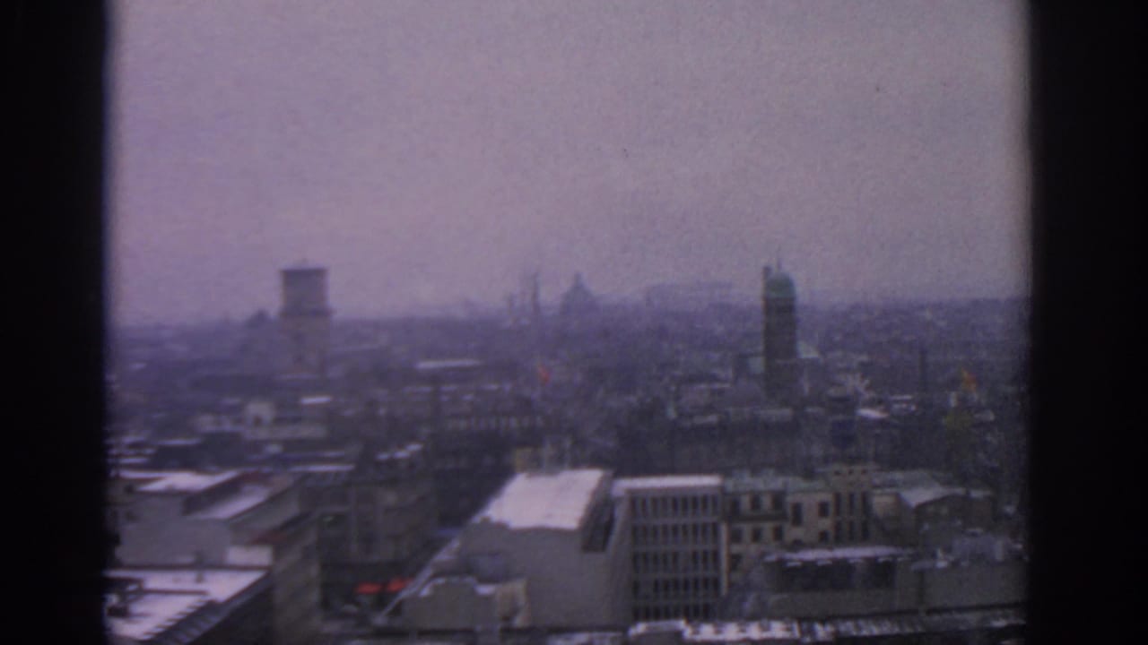 1963 Warsaw city view through a dark window frame, with hazy buildings, rooftop snow, and distant towers under grey sky.