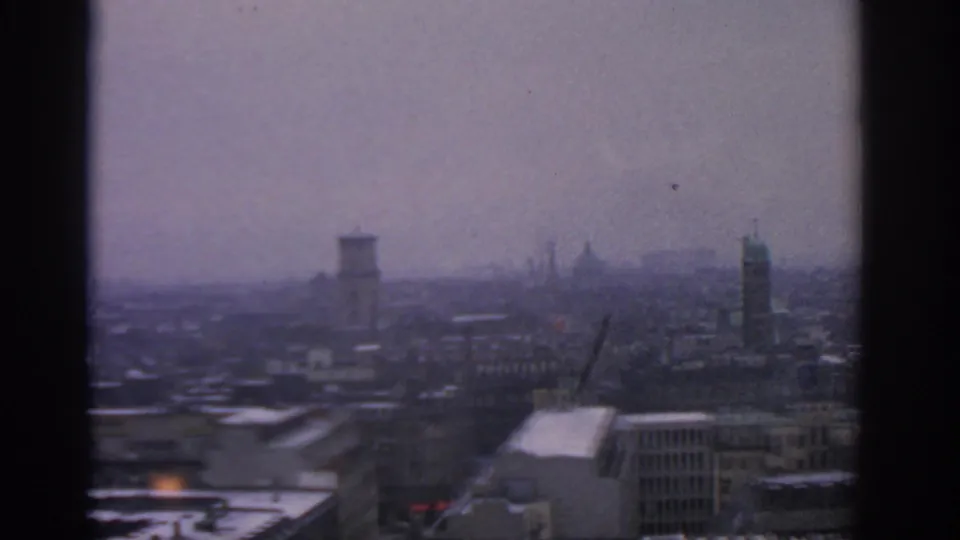 High view across 1963 Warsaw, showing muted grey buildings, distant spires, and a dark vertical window division.