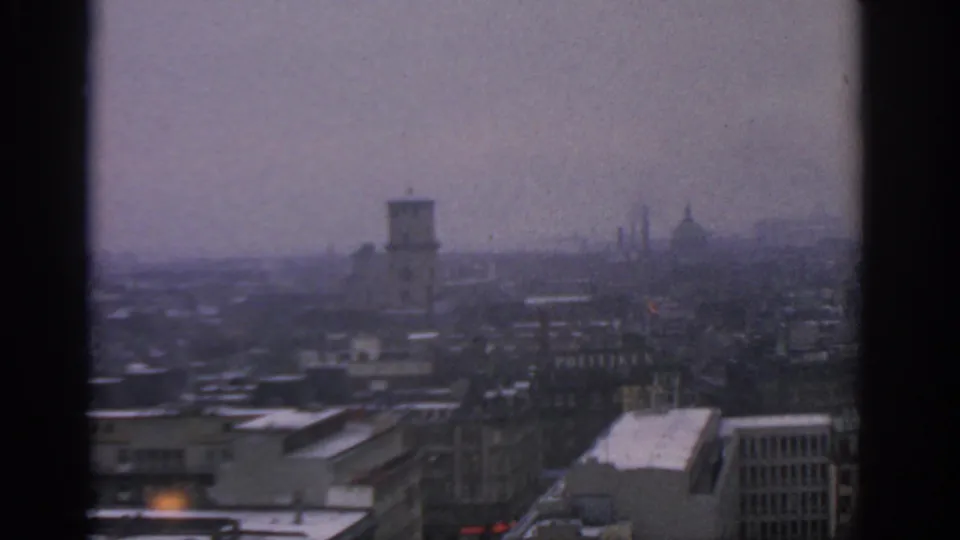 Soft 8mm frame of Warsaw in 1963, with snow on low rooftops, dense buildings, and faint spires in the haze.