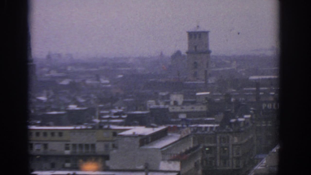 Hazy urban skyline with a tower, snow patches on roofs, and partial sign lettering.