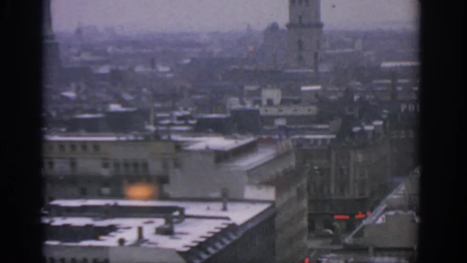 Soft 1963 Warsaw city view with snow-dusted roofs, a tower partly near the top edge, distant spires, and dark 8mm frame borders.