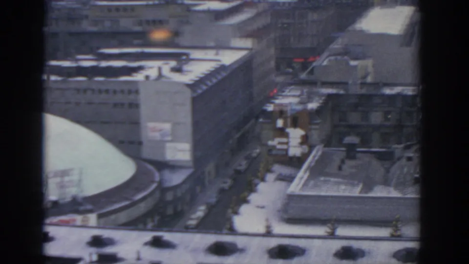 1963 Warsaw elevated frame with snow on rooftops, a narrow street, parked cars, large buildings, and tiny pedestrian figures.