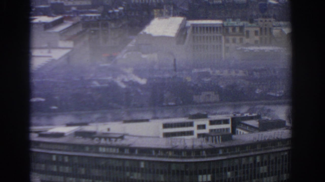 1963 Warsaw cityscape seen from above through dark dividers, with rooftops, a river, smoke plumes, and a grey hazy sky.