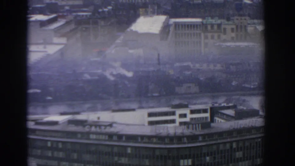 1963 Warsaw cityscape seen from above through dark dividers, with rooftops, a river, smoke plumes, and a grey hazy sky.
