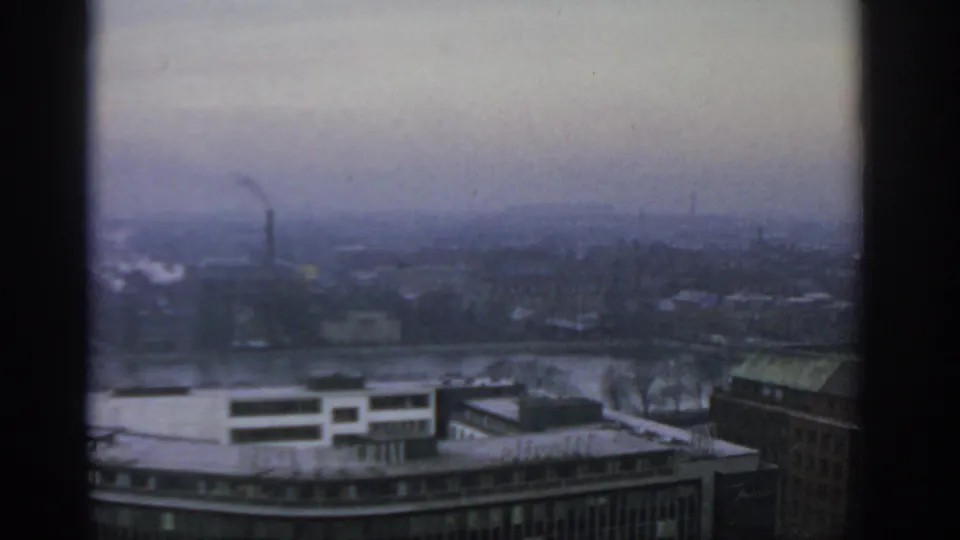 Warsaw in 1963 viewed from a high vantage point, with riverfront buildings, industrial smoke, and a muted grey-blue sky.