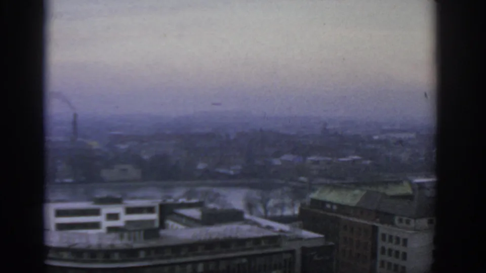 Wide 1963 Warsaw panorama with a river crossing the frame, softened rooftops, distant smoke, and grey haze over the city.