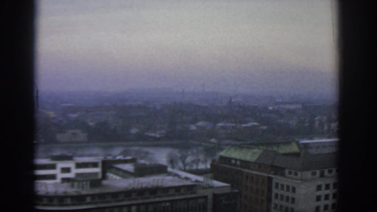 1963 Warsaw home-movie frame showing a hazy elevated city view through a window, with rooftops and a broad waterway visible.