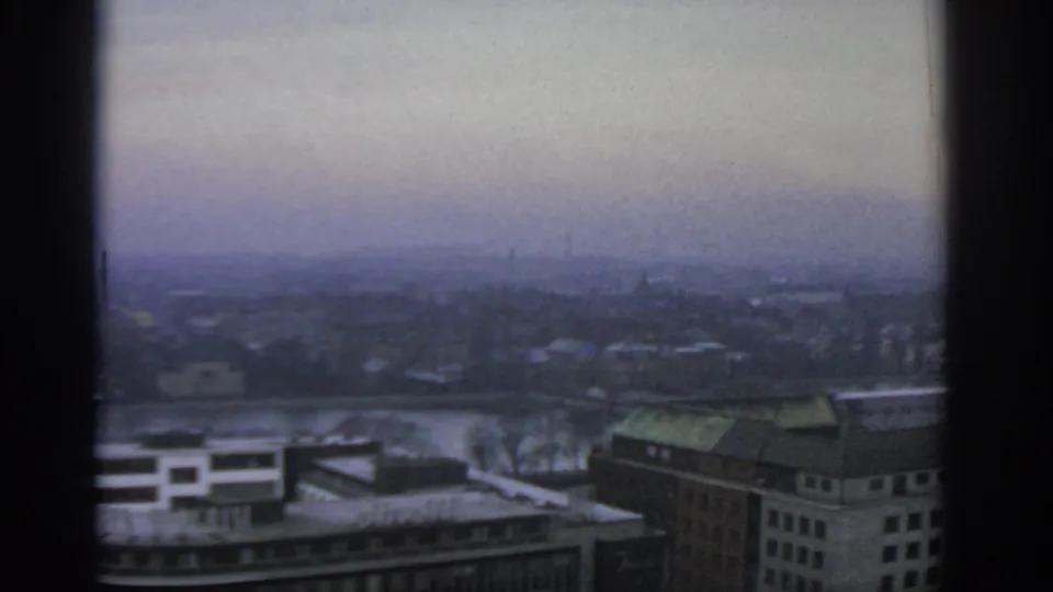 1963 Warsaw home-movie frame showing a hazy elevated city view through a window, with rooftops and a broad waterway visible.