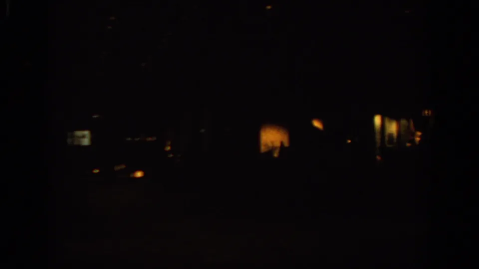 Low-light 8mm frame from the Warsaw travel clip showing scattered warm lights and indistinct shapes inside or beyond a dark interior.