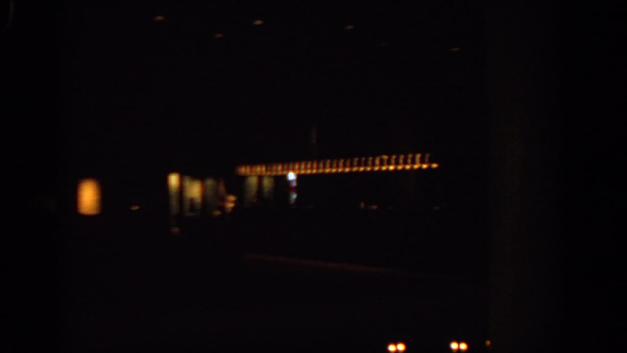Dark moving night view with a row of orange lights and faint building shapes.