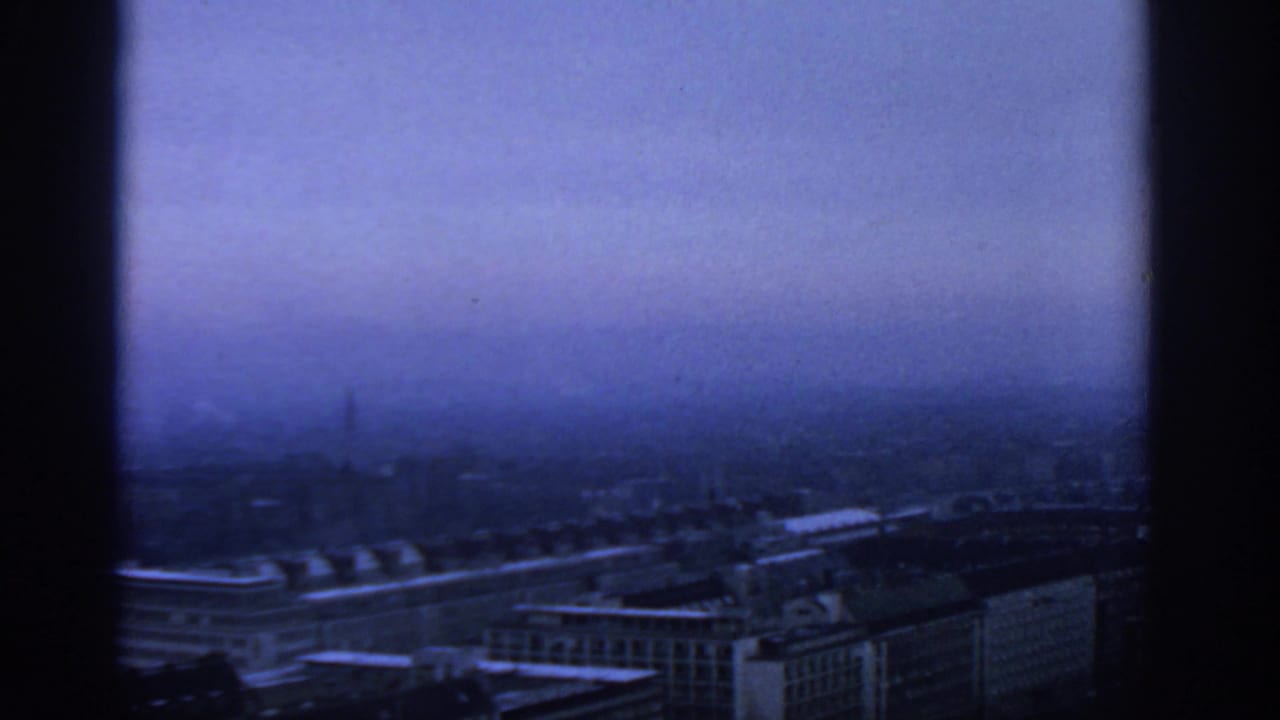 1963 Warsaw skyline appears in blue haze through a high-rise window, with rooftops and distant buildings faintly visible.