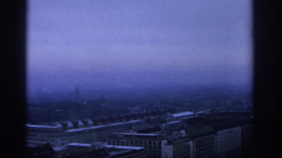 1963 Warsaw skyline appears in blue haze through a high-rise window, with rooftops and distant buildings faintly visible.