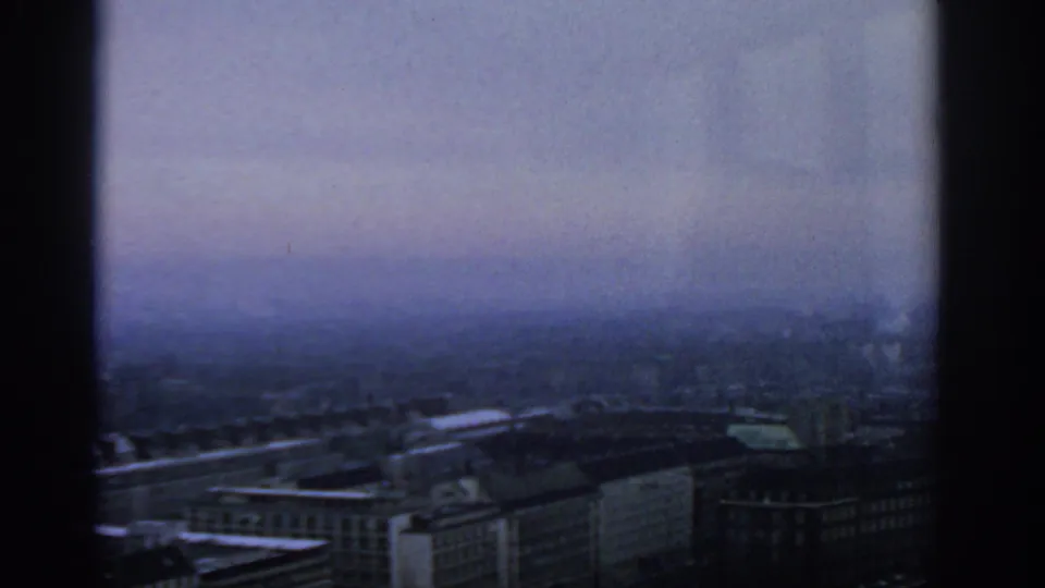 A blue-tinted Warsaw city view shows rooflines and distant urban forms divided by a dark vertical window frame.