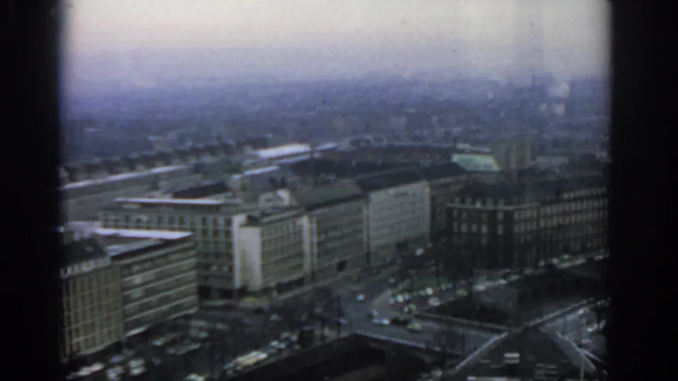 The camera tilts downward over Warsaw buildings, revealing broad streets, rooftops, and parked cars through the window grid.