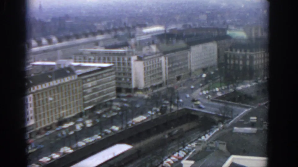 A downward high-rise view over Warsaw shows blocky buildings, a broad street, parked cars, and dark window framing.