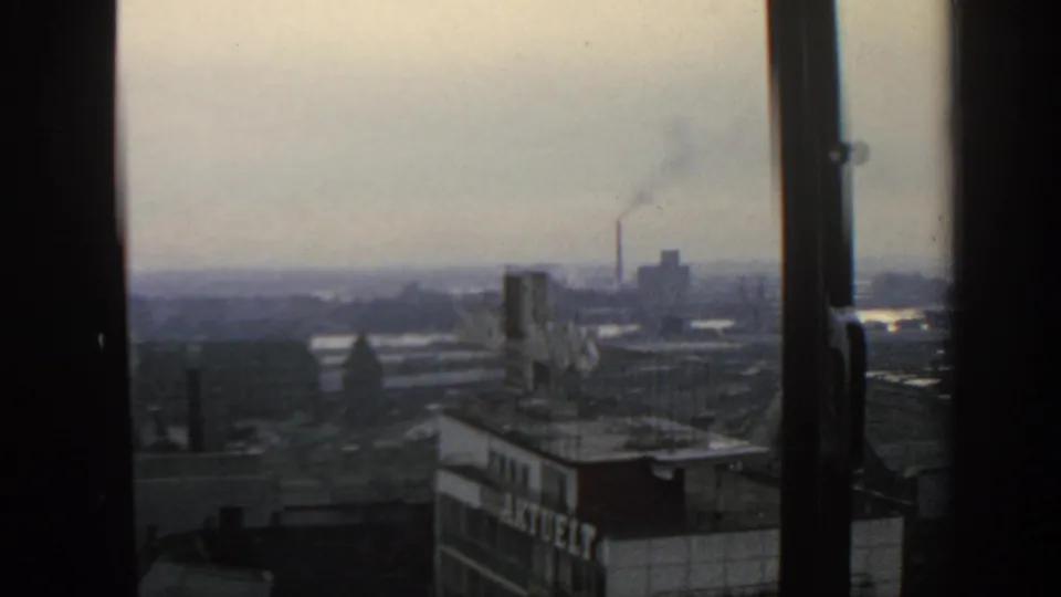 1963 Warsaw skyline through a window, showing rooftops, haze, and an industrial chimney releasing smoke.