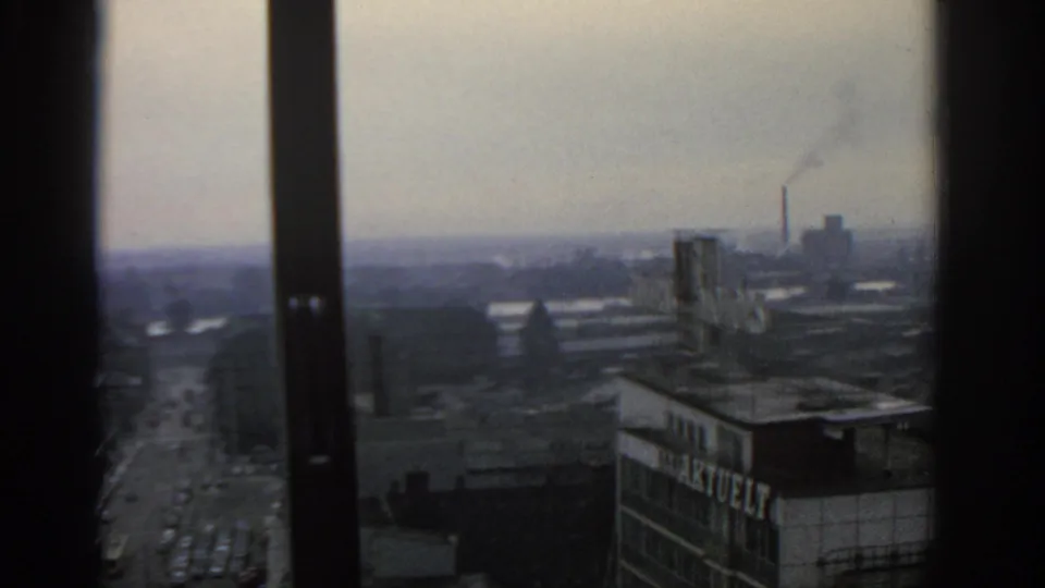 1963 Warsaw cityscape framed by a window, with rooftops, a distant street, haze, and a smoking industrial chimney.