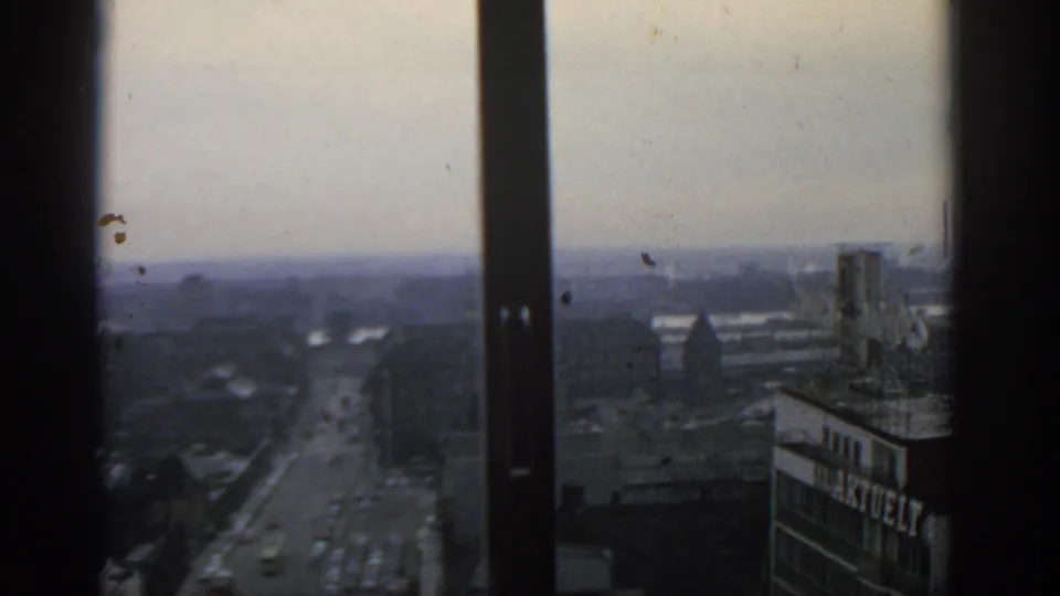 Faded 1963 Warsaw 8mm view through a window, looking over a street, rooftops, haze, and an industrial chimney.