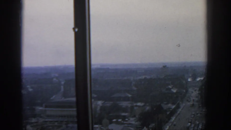 1963 Warsaw cityscape seen from a high window, with dark mullions framing roads, rooftops, and a hazy distant skyline.