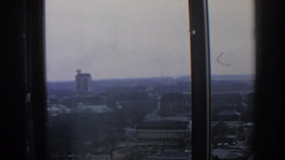 The frame looks across 1963 Warsaw from inside, with a dark window edge, distant tower, low buildings, roads, and muted wintery tones.