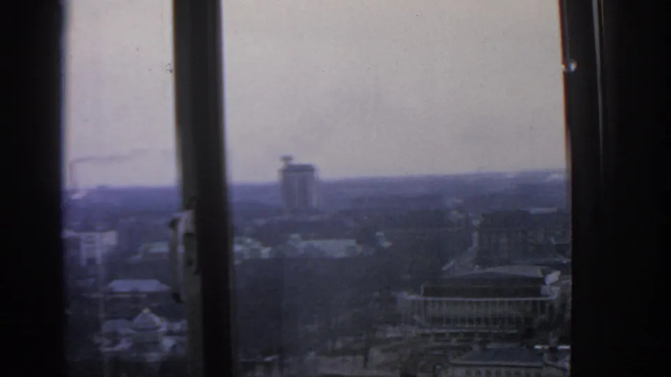 Warsaw appears through multiple dark window mullions, with roads, rooftops, a distant tower, and a pale overcast skyline in 8mm texture.