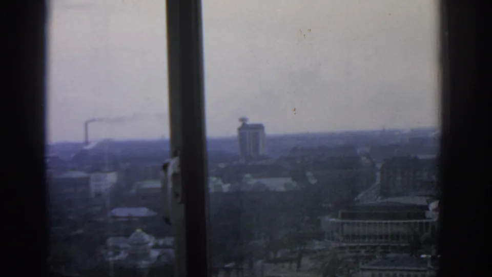 1963 Warsaw cityscape seen through upper-floor window frames, with hazy buildings, bare trees, and smoke on the horizon.