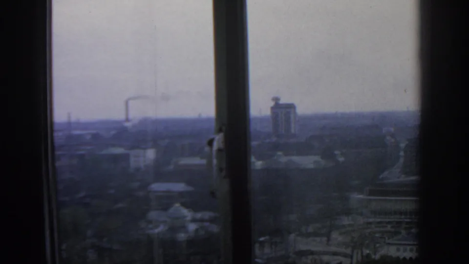 Upper-floor view across Warsaw in 1963, showing a hazy skyline, a tall block-like building, and smoke beyond the window glass.
