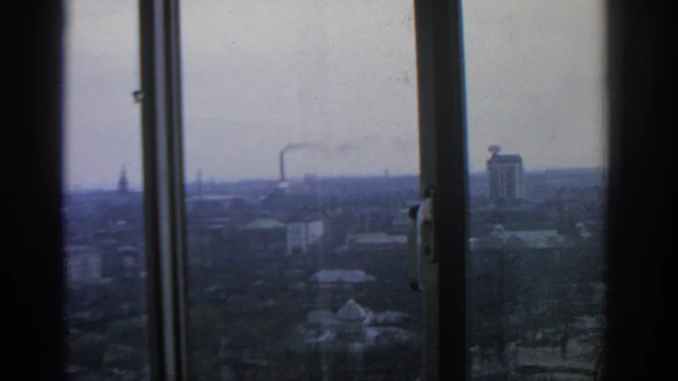 A 1963 Warsaw upper-floor view shows window hardware in the foreground, a hazy cityscape, distant smoke, rooftops, and a slender spire.