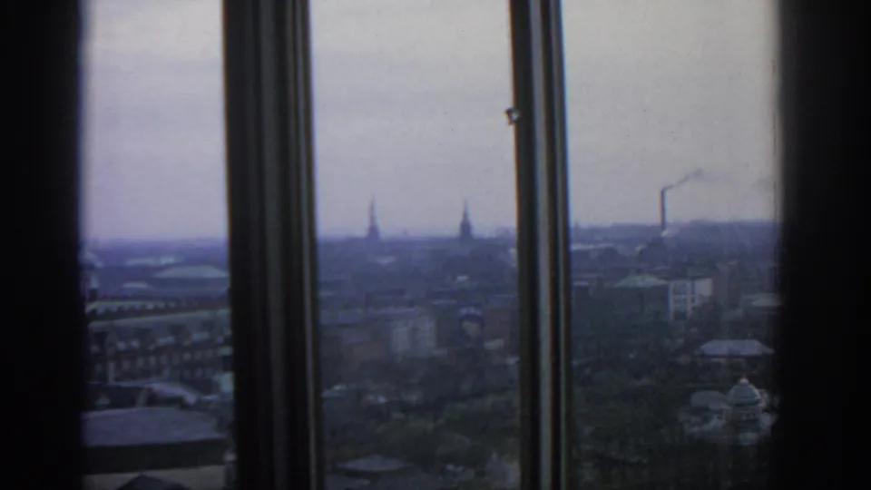 Warsaw cityscape in 1963 seen through tall window panes, with rooftops, spires, and a pale cloudy sky.