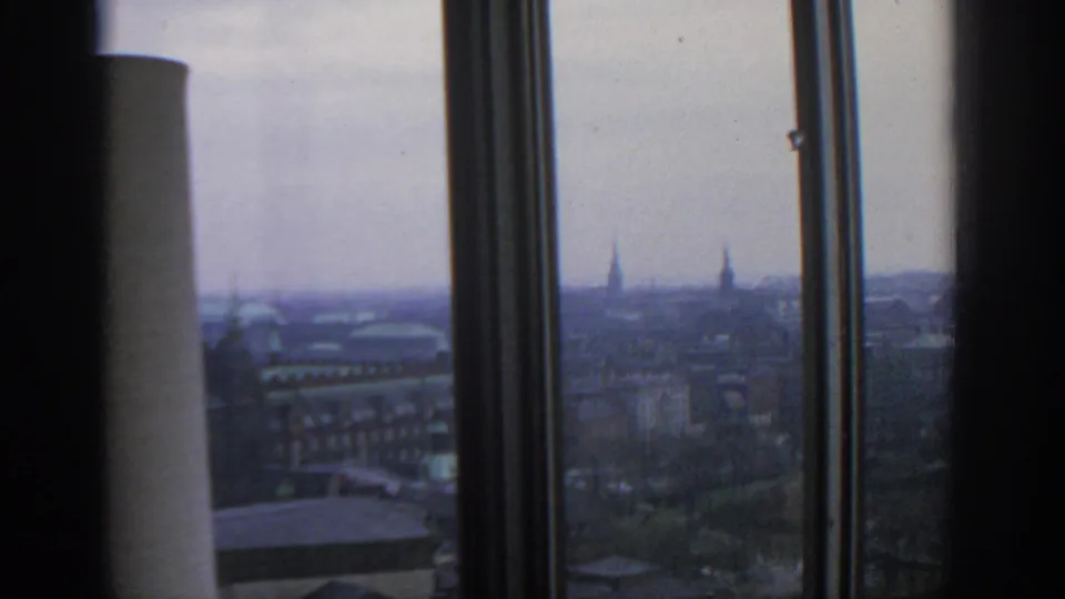 Elevated 1963 Warsaw view through a window, showing layered rooftops, distant spires, and a partial interior column.