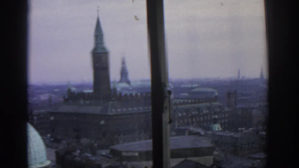 1963 Warsaw skyline through a window, with a prominent clock tower, distant spires, rooftops, and muted overcast light.