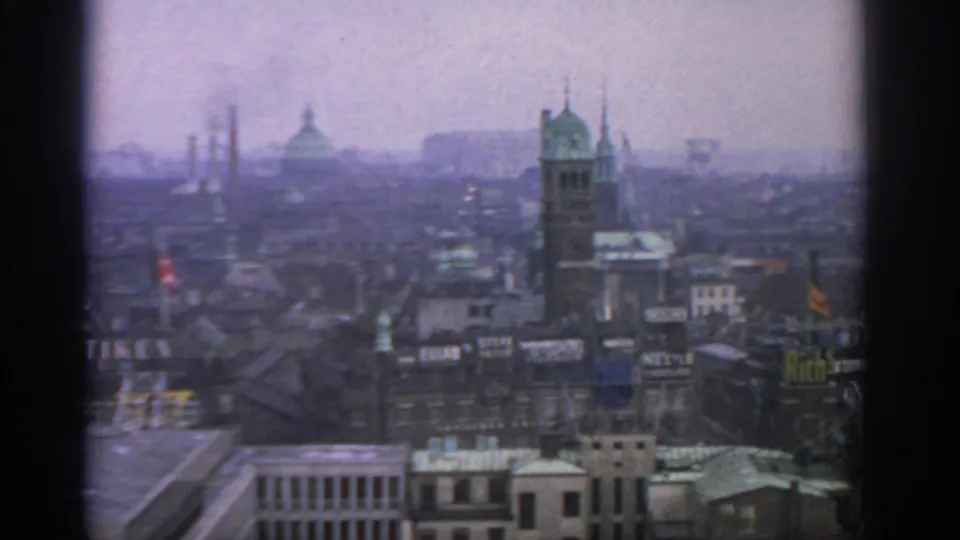 Elevated 1963 Warsaw skyline frame showing a tower, distant dome, smoking chimneys, red flag, and commercial rooftop signs.