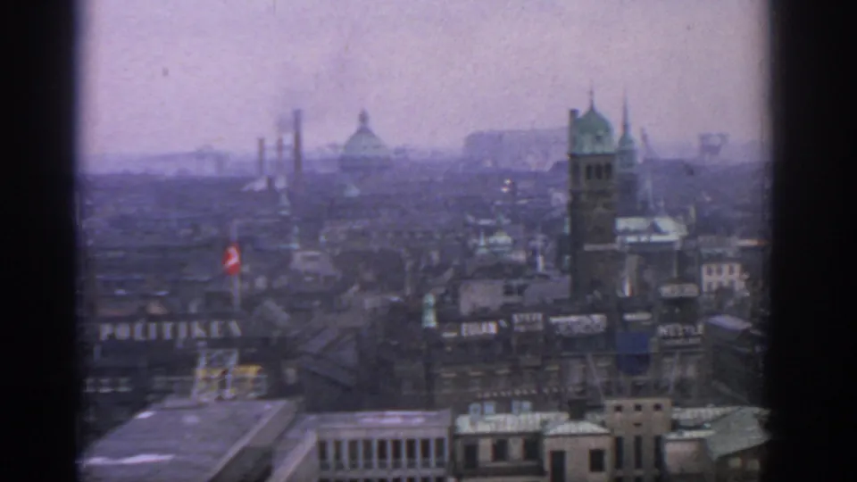 1963 Warsaw skyline seen through 8mm grain, with industrial stacks, a domed roof, a spired tower, and a large visible rooftop sign.