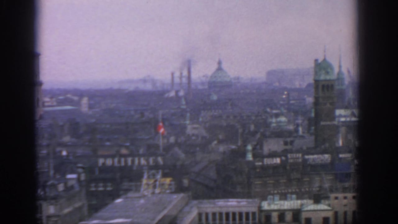 1963 Warsaw aerial view with a hazy skyline, domed buildings, a tower, rooftops, and visible signage through soft 8mm grain.