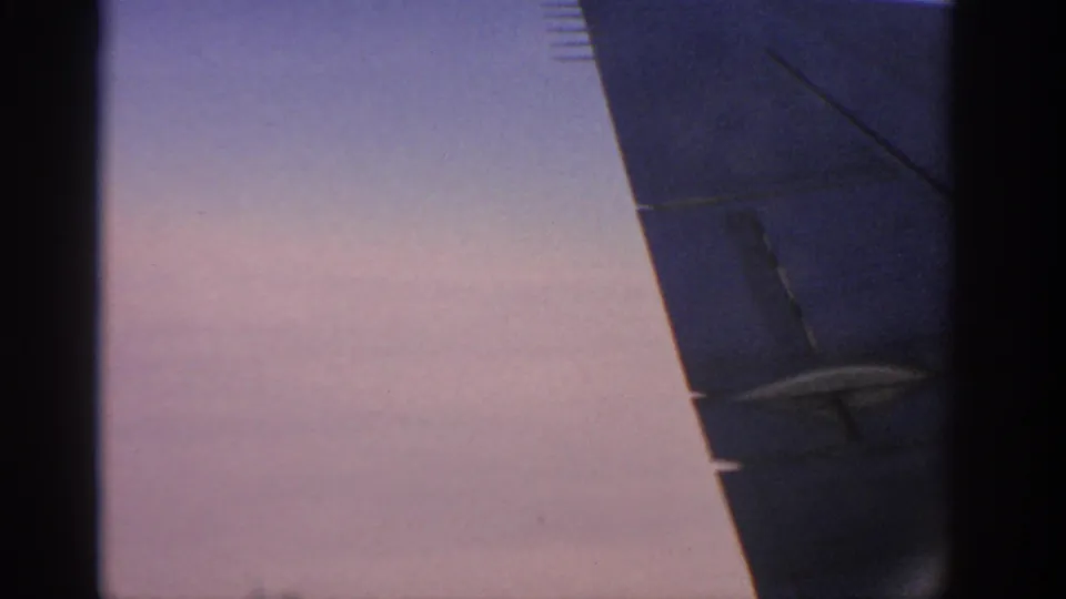 Passenger-plane view with a broad aircraft wing and dark structure silhouetted against a pale sky in 1963 travel footage.