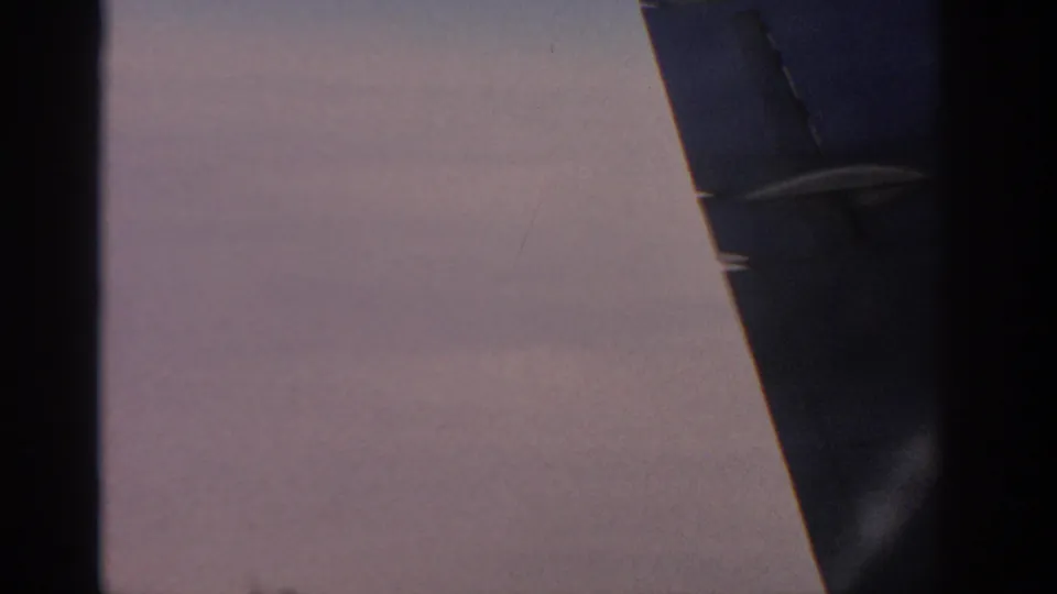 Soft 8mm frame showing an aircraft wing from a passenger viewpoint, with dark plane structure and hazy light beyond.
