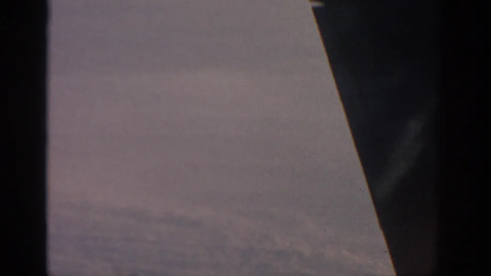 In-flight 1963 view past the aircraft wing, with dark plane structure and faint hazy texture below the soft sky.