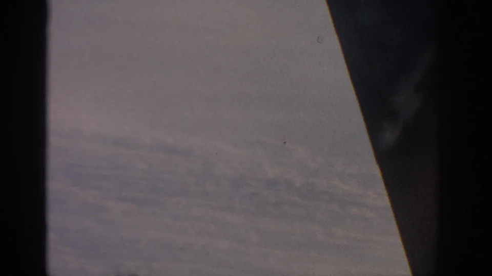 1963 8mm passenger-window view over clouds, framed by dark aircraft or window edges during flight near Warsaw, Poland.