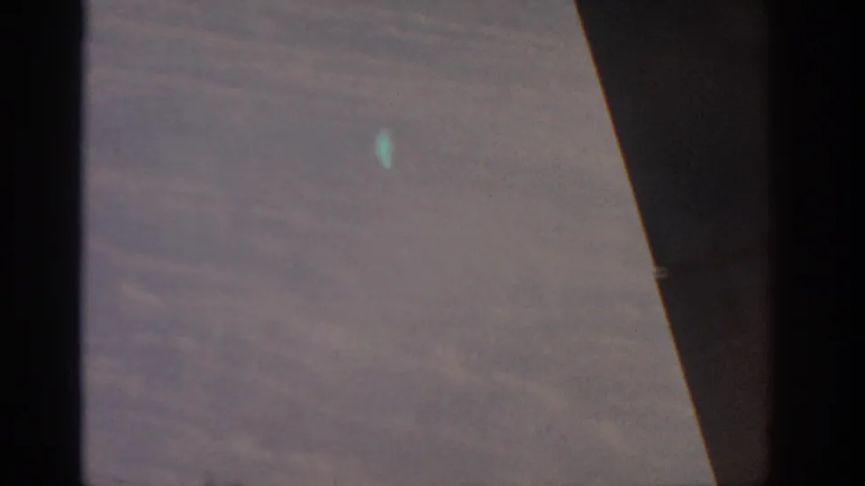 Color 8mm aerial view from inside an aircraft, showing clouds below and a dark wing or fuselage shape in the foreground.