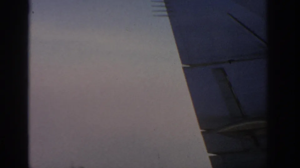 1963 8mm airplane-window view with a visible wing section, panel lines, dark cabin edge, and clouds below.