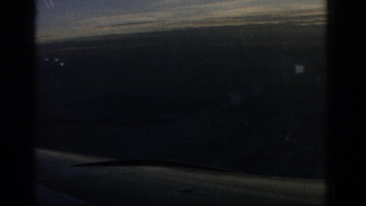 1963 8mm airplane-window view over open water, with a dark window frame, hazy horizon, and aircraft wing edge visible.