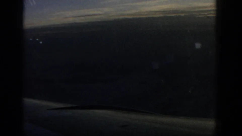 1963 8mm airplane-window view over open water, with a dark window frame, hazy horizon, and aircraft wing edge visible.