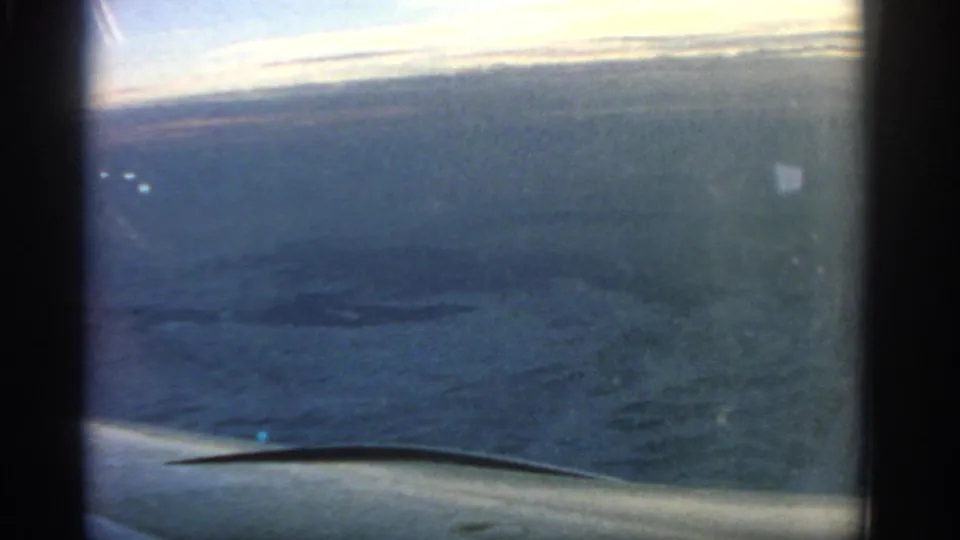 1963 8mm travel frame showing open sea below, a pale horizon, and the airplane wing edge through the passenger window.