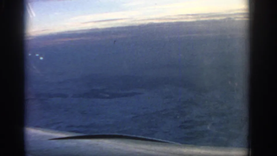 1963 8mm passenger view over the sea, with the aircraft wing edge in the foreground and a bright, hazy horizon above.