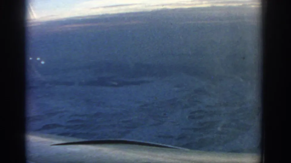 1963 8mm frame from an airplane window, showing textured water below, a soft horizon, and the wing edge in the foreground.