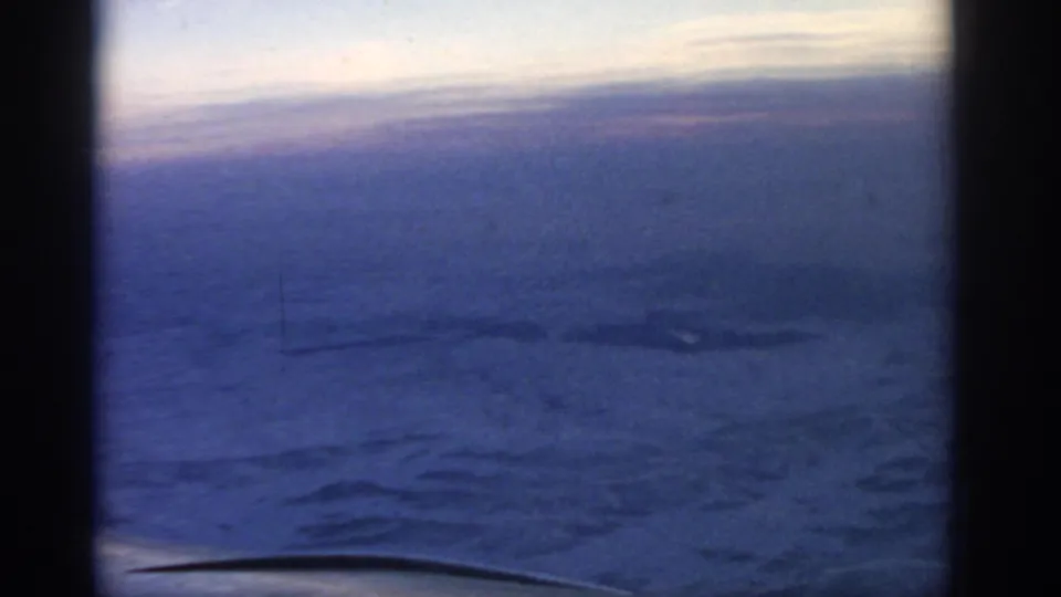 1963 8mm home-movie view from an airplane window, with blue dusk sky, distant horizon, and pale ground far below.