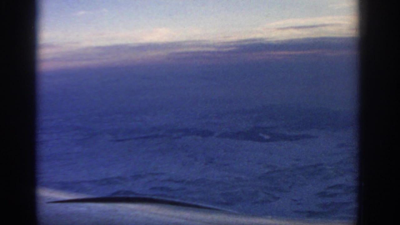 1963 8mm passenger-window view showing an airplane wing over clouds and dim blue-purple sky, with dark window-frame edges.