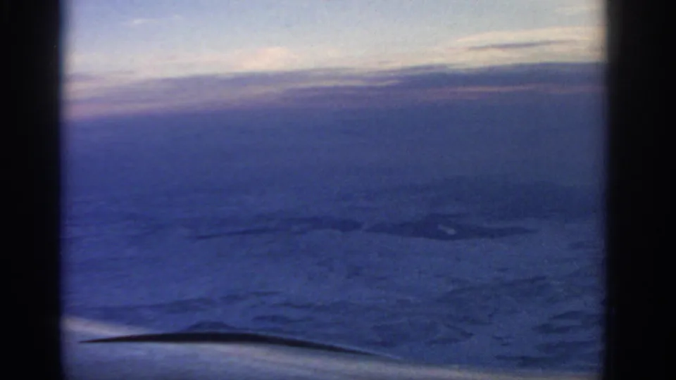 1963 8mm passenger-window view showing an airplane wing over clouds and dim blue-purple sky, with dark window-frame edges.