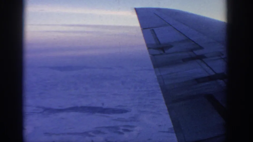 1963 8mm aerial view from an airplane window, with the wing at right and pale cloud layers stretching toward the horizon.