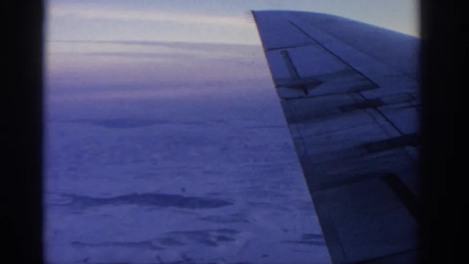 Passenger-window view on 8mm film showing the airplane wing, clouded landscape below, and blue-purple sky during flight.
